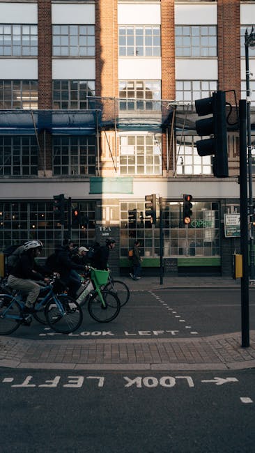 Photograph of a city street in front of a brick building with large glass windows, showing a loading bay area with scaffolding and safety barriers. Below the building, there are traffic lights, a pedestrian crossing, and a street sign indicating 'LOOK LEFT.' Several people are riding bicycles along the street, some wearing helmets. The scene captures an urban environment during daylight, with natural light illuminating the scene. This image relates to house removals and furniture transport, illustrating the logistical considerations of loading and unloading in a busy city setting, as handled by Man with Van Kew in their home relocation and moving services.