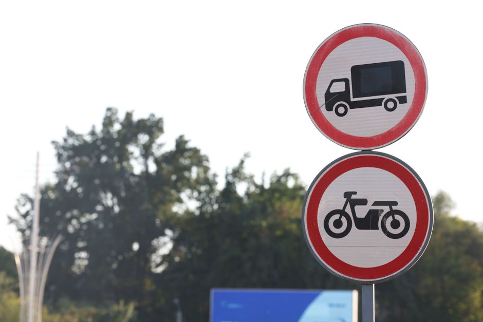 Two circular road signs mounted on a pole outdoors, with a background of trees and a clear sky. The top sign Indicates no entry for large vehicles such as trucks or vans, featuring a black truck icon within a red circle. The bottom sign shows no access for bicycles, with a black motorcycle or bicycle icon inside a red circle. The signs are positioned near a property or road, which could relate to a property undergoing home relocation or furniture transport. The setting suggests a street or loading bay area possibly impacted by local load restrictions or parking regulations relevant to moving services, as provided by Man with Van Kew. This visual detail highlights the importance of understanding loading bay rules and restrictions during the logistics of packing and moving items for house removals.