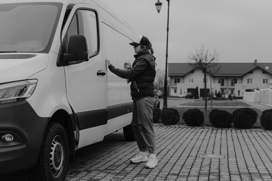 A person dressed in a black puffer jacket, jeans, and white sneakers is seen stepping out of a white van with a black side panel, parked on a cobblestone street. The individual's face is not visible a