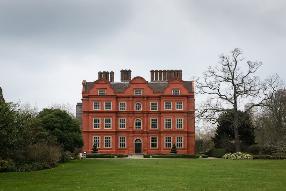 A large, historic red-brick building with multiple windows and decorative architectural details, situated on a well-maintained grassy lawn with trimmed shrubs and bushes. The scene shows an overcast sky with grey clouds, and leafless trees border the property, indicating a winter or early spring setting. In the foreground, a professional removal team is in the process of handling fragile antiques as part of a home relocation or furniture transport service. The team uses equipment such as cardboard packing materials, moving blankets, and a trolley to carefully position items near the building's entrance. The van is not visible in this shot, but the overall environment suggests organized moving logistics undertaken by [COMPANY_NAME], specialists in house removals and packing and moving services, operating near Kew Gardens, KEW.