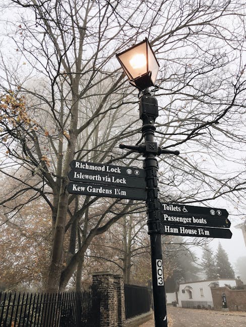 A tall black street lamp with an illuminated lantern at the top standing amidst leafless trees on a foggy day. Attached to the lamp post are multiple black directional signs with white lettering indicating nearby locations and distances, including Richmond Lock, Isleworth via Lock, Kew Gardens 1 mile, Toilets & Passenger boats, and Ham House 1/4 mile. The signs suggest a location close to a historic or scenic area, potentially in or near Kew. Behind the lamp post, a brick fence and a small white building are partially visible through the haze. The scene reflects a quiet, outdoor environment suitable for home relocation or moving services, with the signs providing contextual clues for navigation around the area, relevant to the process of packing and transporting belongings during house removals in the vicinity of Kew Green.