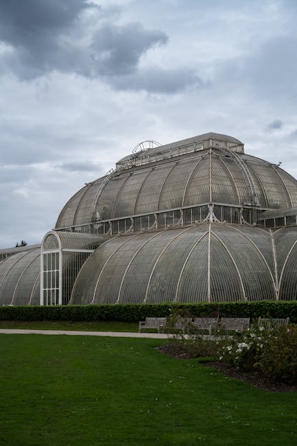 The image depicts a large Victorian-style glass greenhouse with multiple curved glass panels and metal framing. The structure is situated outdoors on a well-maintained lawn with neatly trimmed bushes and a flower bed in the foreground. The greenhouse features a prominent central dome with a rounded skylight at the top, and an arched window on the side. The overcast sky above has dark grey clouds, creating a subdued lighting environment. This type of building is commonly associated with historic botanical gardens or conservatories, which may require careful relocation during house removals. Man with Van Kew, a professional moving service, could assist with transporting such delicate and substantial glass structures during home relocation or furniture transport processes. The scene emphasizes the importance of strategic packing, handling fragile materials, and meticulous loading procedures involved in moving architectural glass conservatories, especially in tight-access Victorian homes.