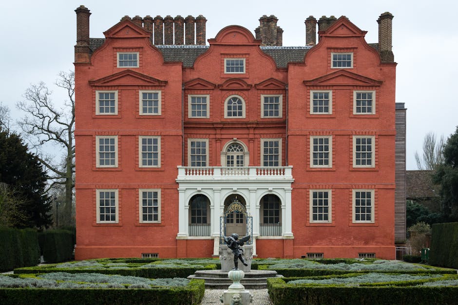 A tall black street lamp with an illuminated lantern at the top standing amidst leafless trees on a foggy day. Attached to the lamp post are multiple black directional signs with white lettering indicating nearby locations and distances, including Richmond Lock, Isleworth via Lock, Kew Gardens 1 mile, Toilets & Passenger boats, and Ham House 1/4 mile. The signs suggest a location close to a historic or scenic area, potentially in or near Kew. Behind the lamp post, a brick fence and a small white building are partially visible through the haze. The scene reflects a quiet, outdoor environment suitable for home relocation or moving services, with the signs providing contextual clues for navigation around the area, relevant to the process of packing and transporting belongings during house removals in the vicinity of Kew Green.