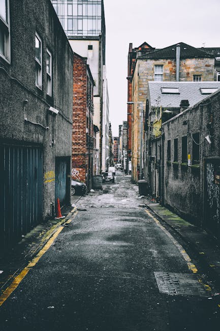A narrow urban alleyway in Kew's Sandycombe Road featuring a wet, dark asphalt surface with yellow line markers along the edges, flanked by tall, weathered brick and concrete buildings on both sides. The left side has a blue metal garage door, while the right side displays a high concrete wall with a small yellow post and some graffiti. In the background, a white van is visible parked further down the alley, with a few residential buildings and rooftops extending into a cloudy sky. The setting suggests a typical city street scene suited for house removals or furniture transport, with no people or moving equipment present in the visible shot. Man with Van Kew specialises in home relocation services, including parking and access planning for narrow streets like Sandycombe Road, as part of their removals operations.