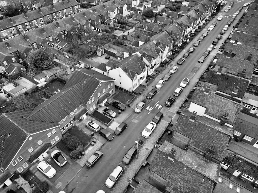 An aerial black-and-white photograph of Sandycombe Road in Kew showing a narrow residential street lined with closely spaced terraced houses and semi-detached homes. Cars are parked along both sides of the street, with some parked on the pavement, and others aligned parallel to the curb. A parked van, equipped with loading straps and covered in protective blankets, is positioned near the pavement, indicating an ongoing home relocation or furniture transport process. The image captures the residential environment, including rooftops, small gardens, and narrow pathways between properties, emphasizing the constraints of moving through such tightly packed streets. The scene reflects the logistical considerations for house removals in dense urban areas, with a focus on careful loading and transportation of household items by a professional removal company such as Man with Van Kew, ensuring efficient packing and safe vehicle loading within the limited space.
