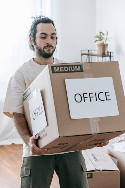 A man with dark, curly hair and a beard is inside a bright, modern room, carrying a large brown cardboard box labeled 'OFFICE' with a smaller label indicating 'MEDIUM' size. The box is securely sealed