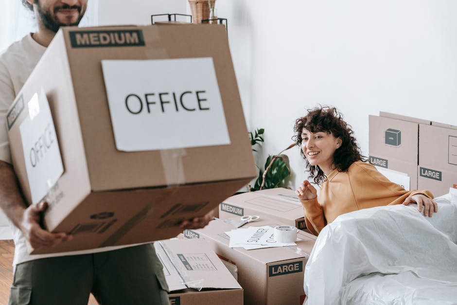 A man with dark, curly hair and a beard is inside a bright, modern room, carrying a large brown cardboard box labeled 'OFFICE' with a smaller label indicating 'MEDIUM' size. The box is securely sealed