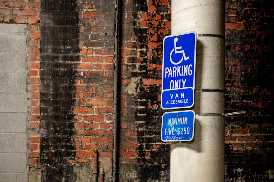 A close-up view of a blue parking sign affixed to a concrete utility pole, indicating designated parking for disabled individuals and van access, with an additional smaller sign below specifying a minimum fine of $250. The background features a weathered brick wall with remnants of black soot and an adjacent concrete wall, suggesting an urban setting. The sign is positioned near a parking area that may be used during house relocation or furniture transport, with the sign serving as a visual cue for accessible parking regulations relevant to professional removal services, such as those provided by Man with Van Kew, operating in the KEW area. The lighting appears natural, and the signage is clearly visible, emphasizing the importance of parking adherence during loading and unloading processes associated with home removals or relocations.
